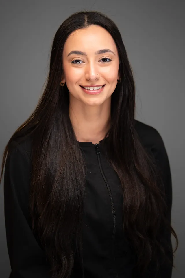 Dental team member with long dark hair in a black jacket, studio headshot on a grey backdrop