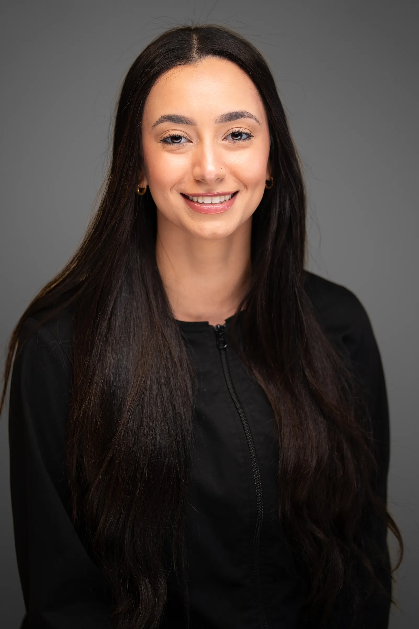 Dental team member with long dark hair in a black jacket, studio headshot on a grey backdrop