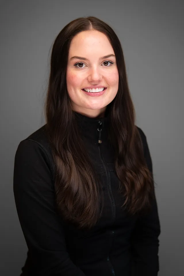 Smiling dental team member in a black zip jacket, studio headshot on a grey backdrop