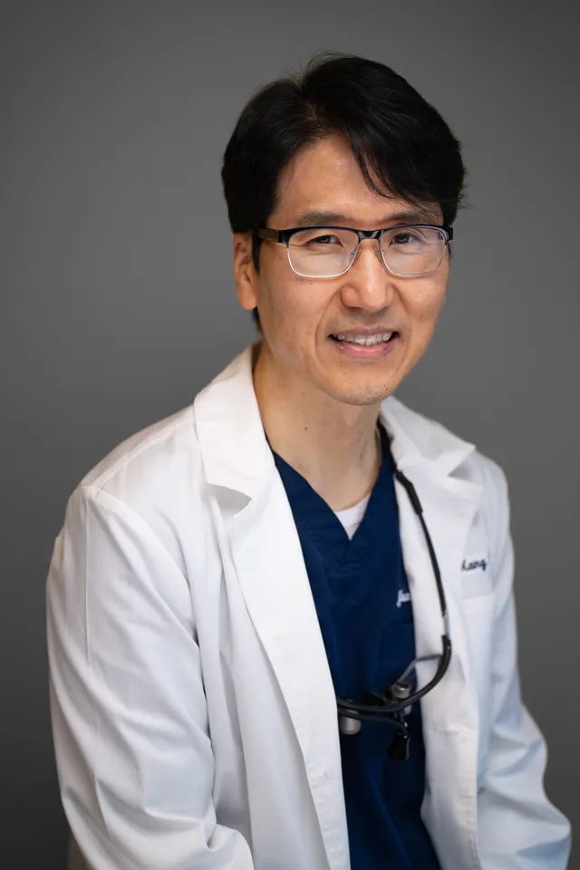 Dentist in a white coat and glasses smiling for a studio headshot on a grey backdrop