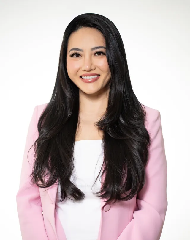 Aesthetic injector in a pink blazer smiling with hands together, on a white backdrop