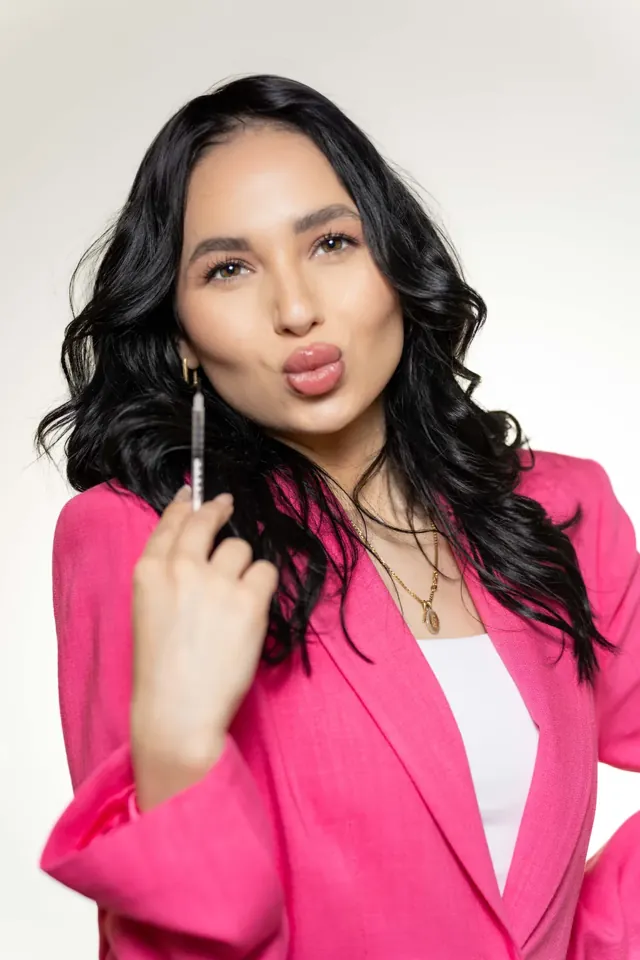 Aesthetic injector in a pink blazer blowing a kiss to the camera while holding up a syringe, vertical branding portrait on a white backdrop