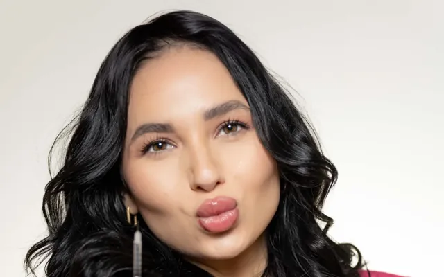 Personal branding portrait of an aesthetic injector with long dark wavy hair and a pink blazer, lips pursed toward the camera against a cream backdrop