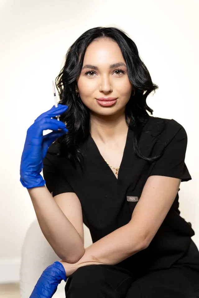 Aesthetic injector in black scrubs seated against a cream backdrop, holding a syringe in a blue gloved hand