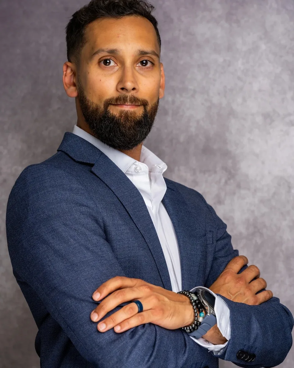 Professional headshot of a man in a navy blazer with arms crossed against a grey studio backdrop