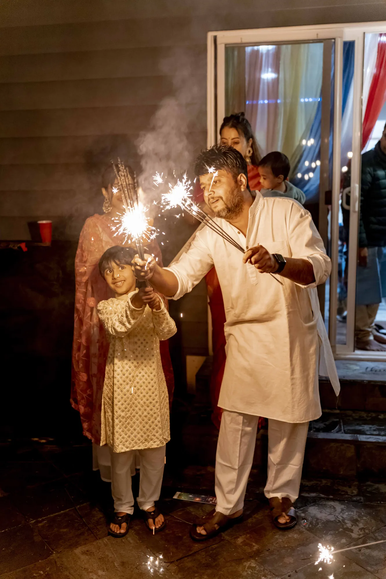 Father and son in white and cream kurtas lighting sparklers together while family watches from a doorway on Diwali night