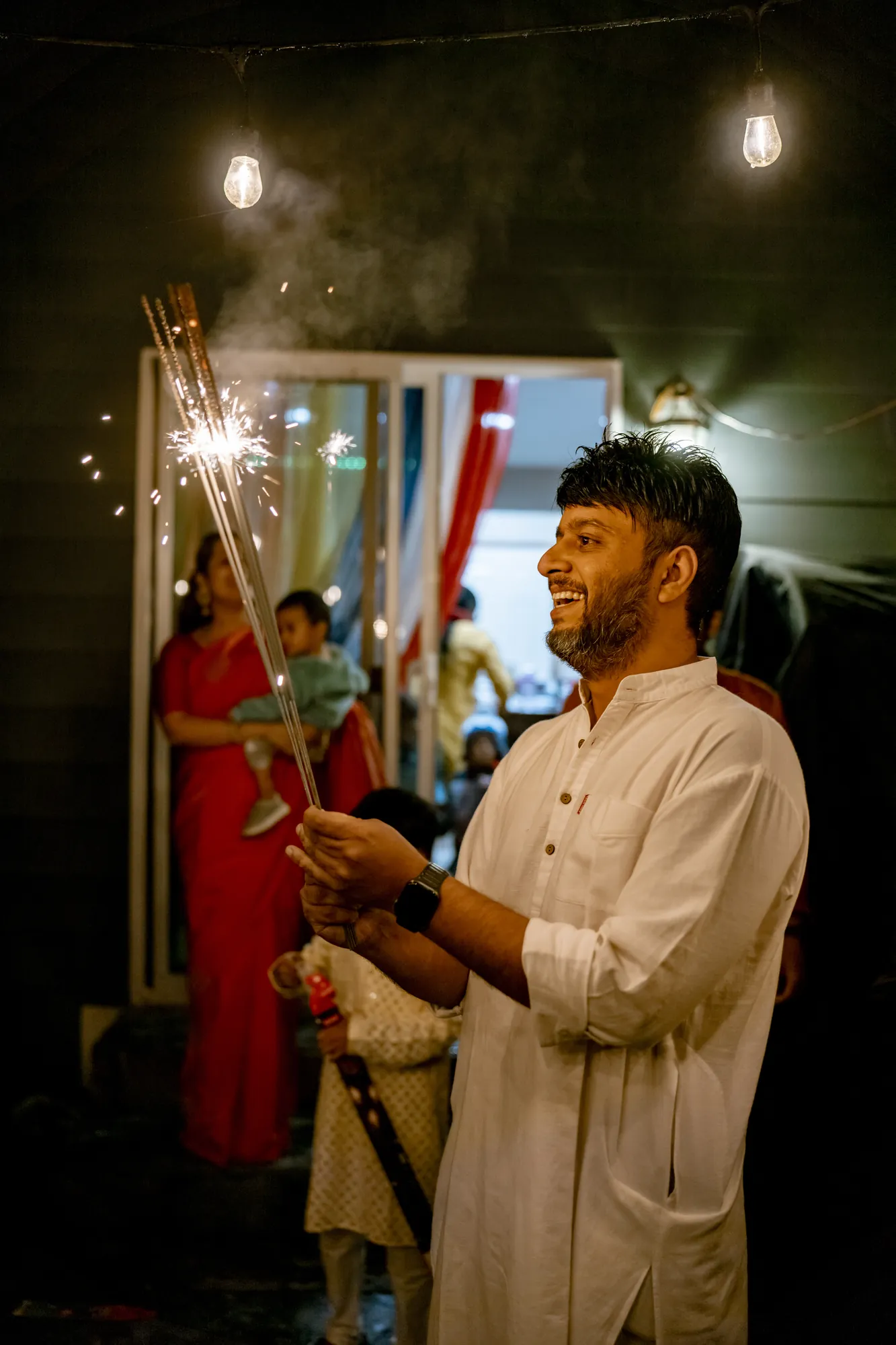 Man in a white kurta laughing as he holds a bundle of lit sparklers under string lights