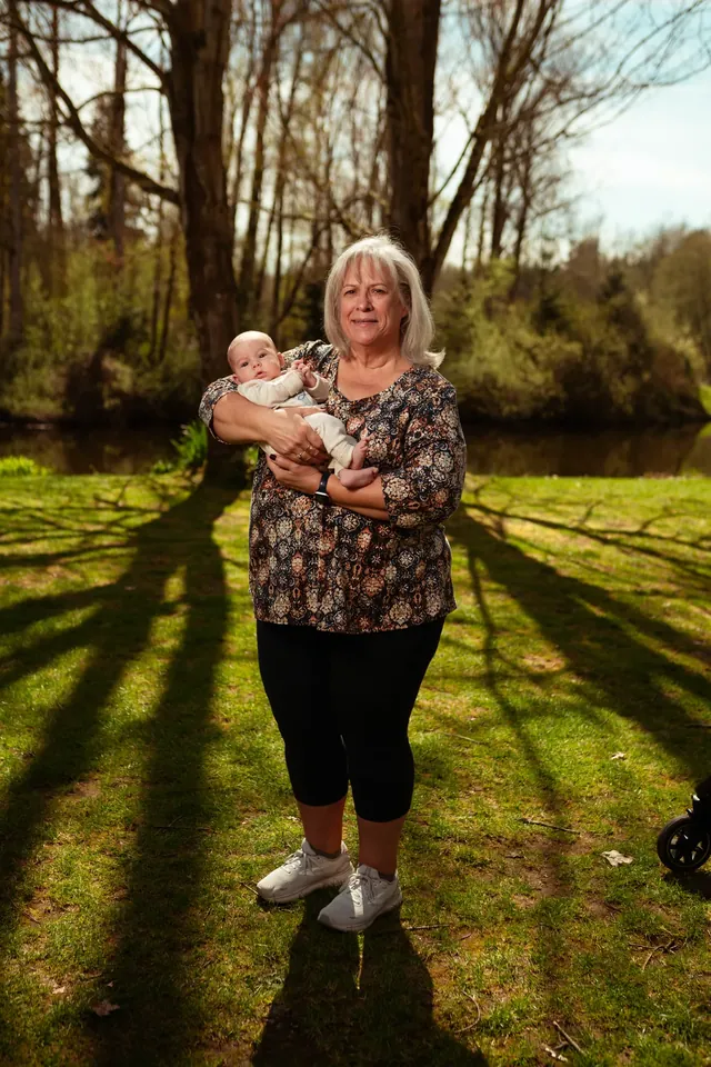 Grandmother with silver hair holding a baby in a cream outfit on a sunny lawn, with a pond and tall bare-branched trees behind them