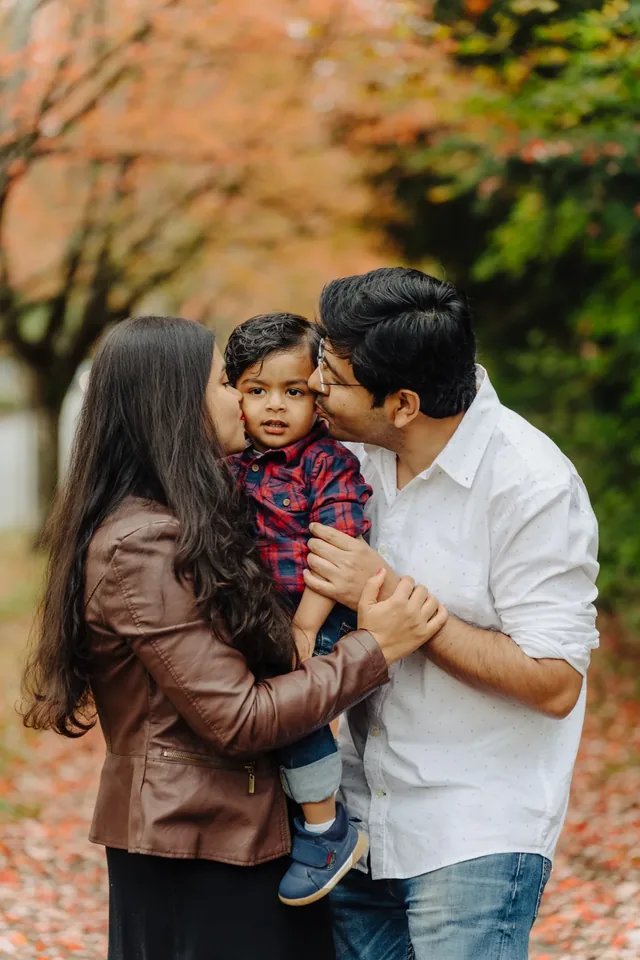 Parents kissing their toddler's cheeks on a leaf-covered autumn path