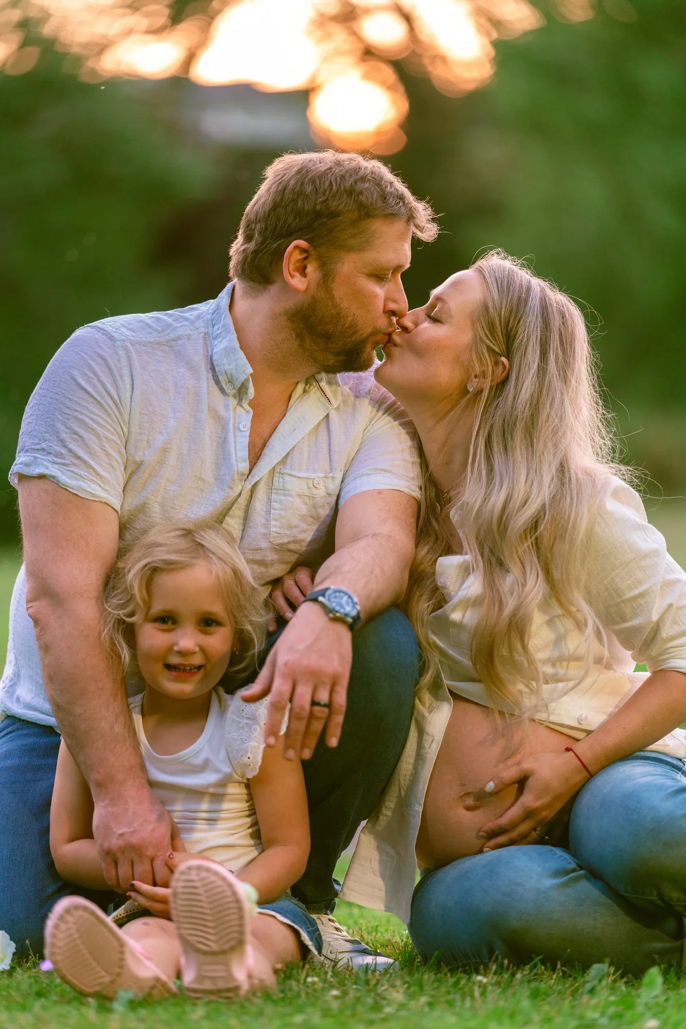 Parents kissing with big sister smiling underneath sunset bokeh at a family maternity session