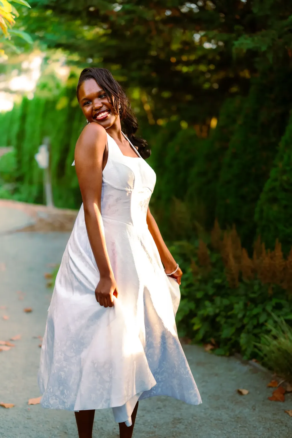 Senior in a white dress glancing back on a garden path in evening light