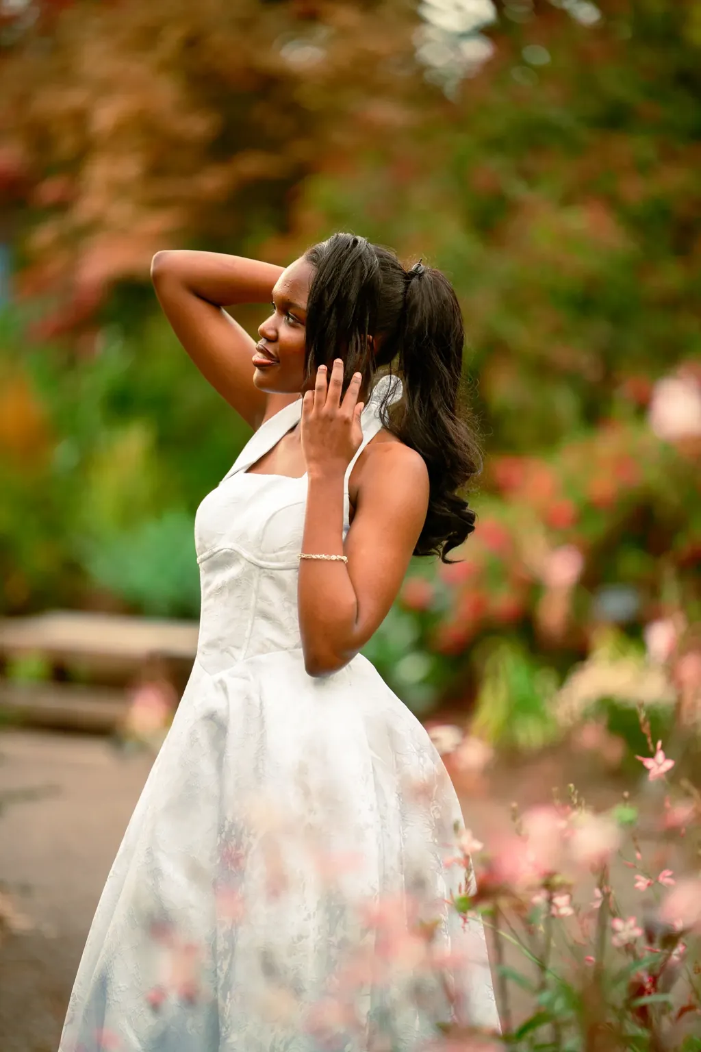 Senior in a white dress with one hand in her hair among garden blooms