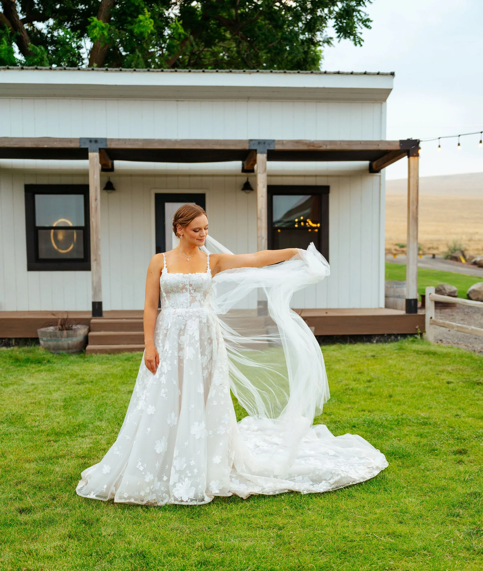 Bride in a floral lace gown outside a white farmhouse, train spread across the grass