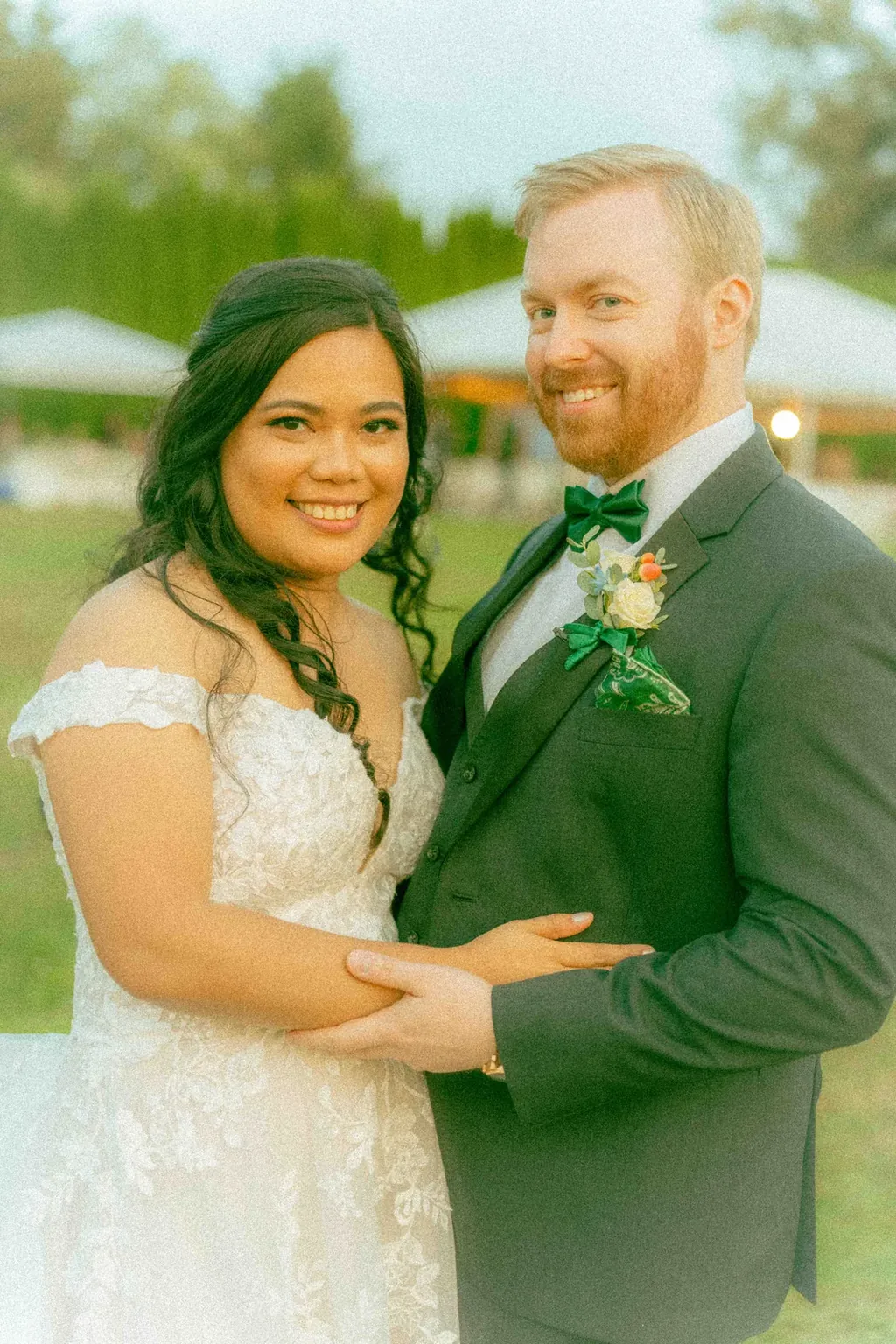 Bride and groom smiling on an open lawn at golden hour, a white reception tent behind them